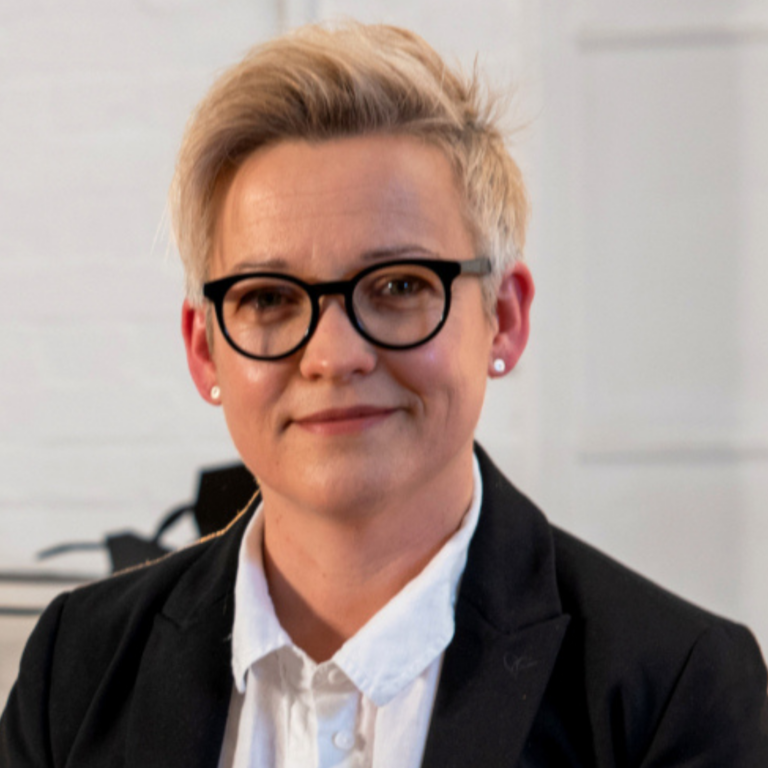 This is a picture of Dr Kate Goodger. She is a white woman with short blond hair and dark-rimmed glasses. Dr Goodger is seated and wearing a white shirt and black jacket.