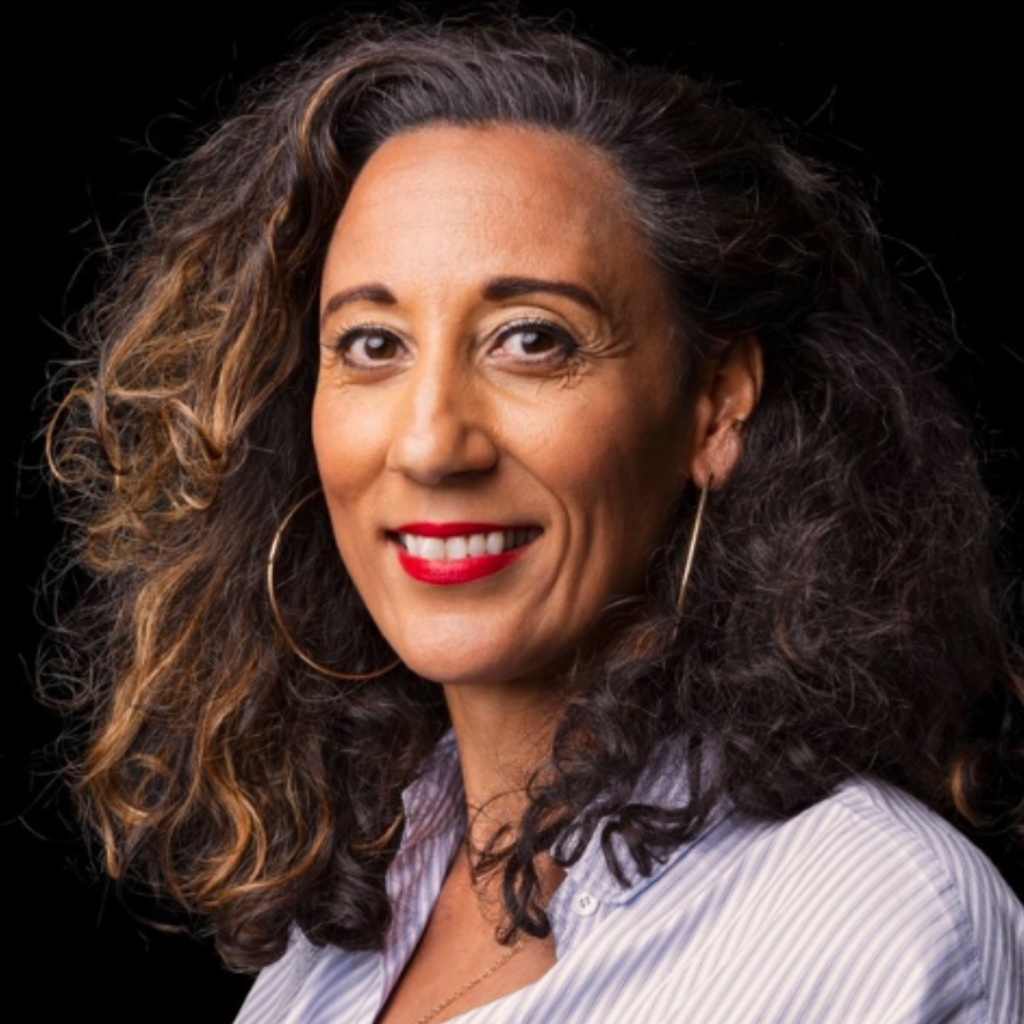 Michelle is a black woman of mixed heritage. She is smiling, and wears red lipstick. The image is set against a dark background. She is wearing a white shirt.