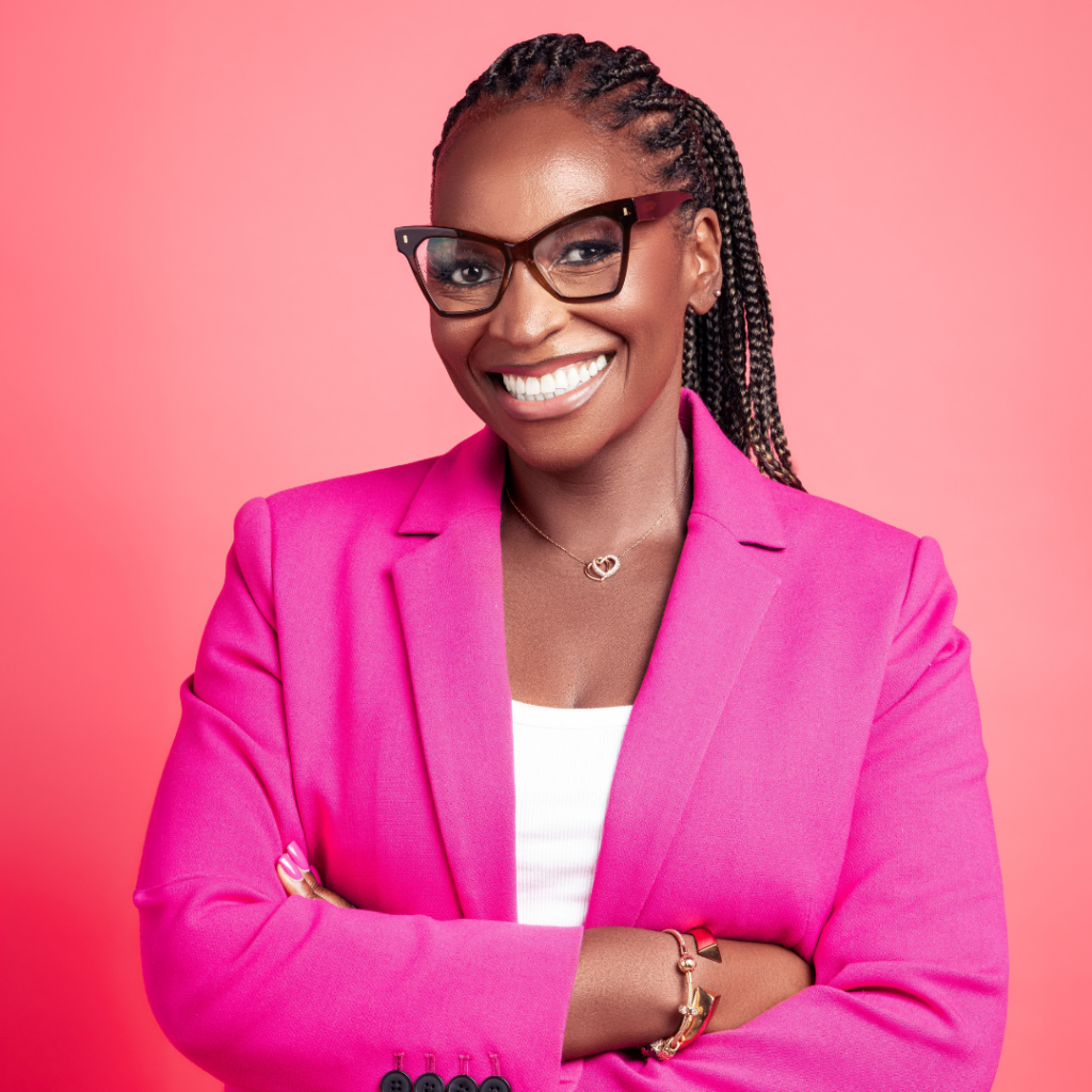 Arit Eminue is wearing a hot pink suit with a white vest. She is standing against a pale pink background. She is smiling and wearing large brown glasses. She has hair long braids pulled into a high ponytail.