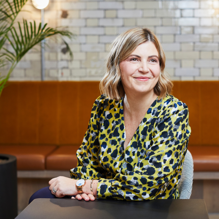 Maike Currie is wearing a floral v-neck blouse. She is a white woman with blond hair. She is sitting at a table and smiling.